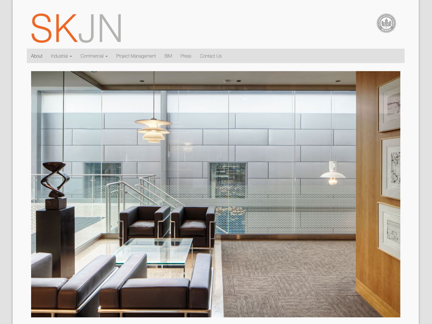 A screen capture of a website page with a clean, white, minimalist design in white and light grey with a simple, straight character font for the title "SKJN", of which the first two letters are orange and the second two grey, below that a navigation bar with some links in grey and across the rest of the page is the masthead photo of a clean, very modern building interior with square-shaped chairs and a wood finish was visible near a metal staircase
